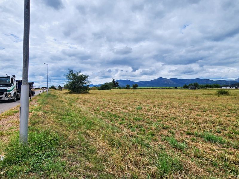 A view of the vacant commercial land parcel from the roadside, featuring a paved verge and open field with mountain views in the distance.