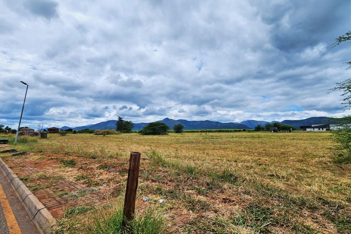 A wide-angle view of a vacant commercial land plot featuring street-side access, paved kerbing, and scenic mountain views in the background.