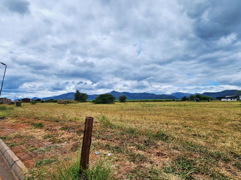 A wide-angle view of a vacant commercial land plot featuring street-side access, paved kerbing, and scenic mountain views in the background.