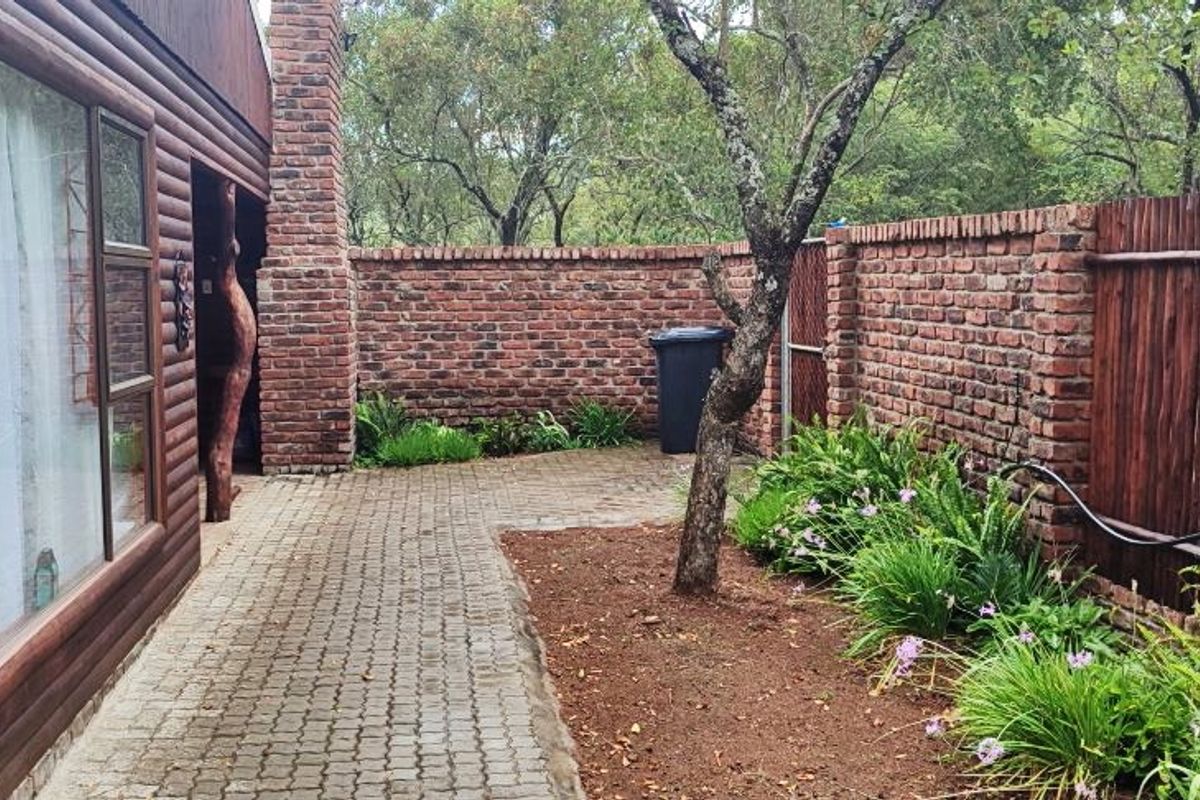 A private rear courtyard featuring a paved walkway, rustic brick boundary walls, and established garden beds adjacent to a timber-clad exterior wall.