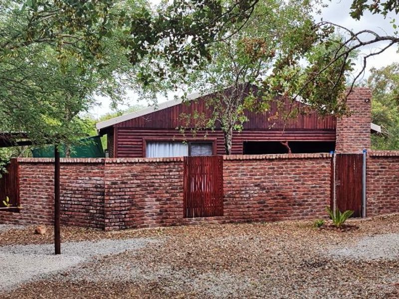 Rear view of a rustic timber-clad house featuring a brick boundary wall, chimney, and a timber pergola set within a natural wooded environment.