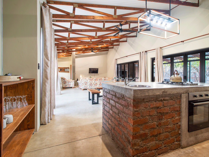 Spacious open-plan living area featuring exposed timber roof trusses, a rustic brick kitchen island with built-in appliances, and large glass doors leading outdoors.
