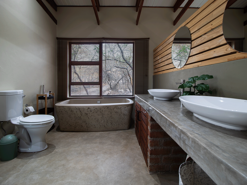 A spacious bathroom featuring a freestanding bathtub, double vessel sinks on a concrete vanity with exposed brick base, and rustic timber ceiling trusses.