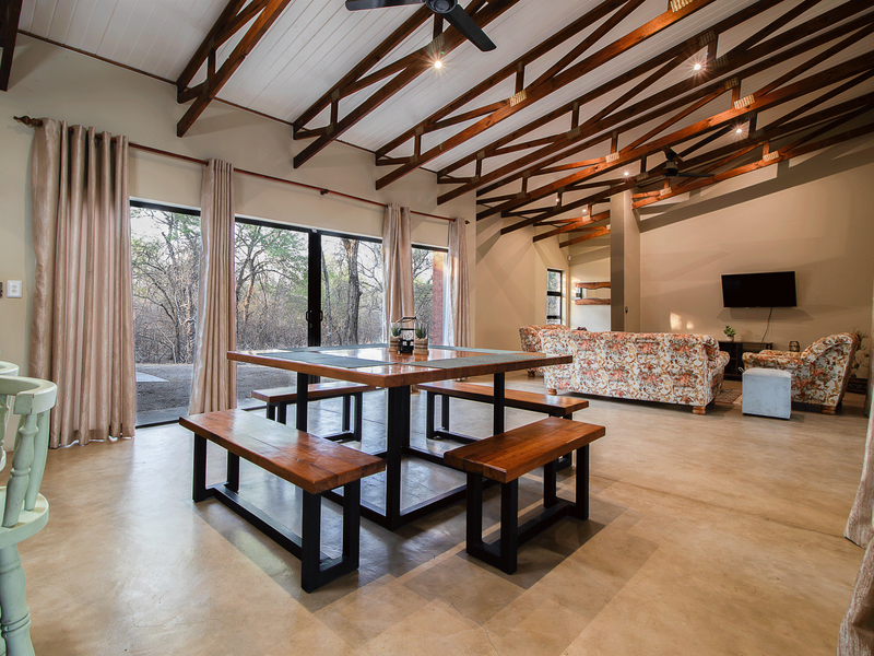 Spacious open-plan living and dining area featuring high vaulted ceilings with exposed timber trusses, polished concrete floors, and large sliding doors leading outdoors.