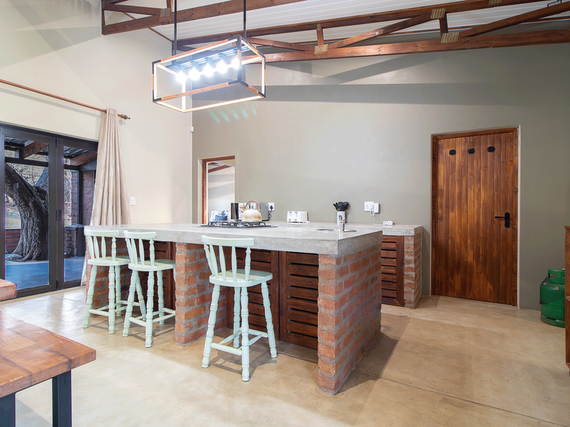 A rustic-style kitchen featuring a large concrete island with a brick base, exposed timber roof trusses, and polished concrete floors, opening to an outdoor area.