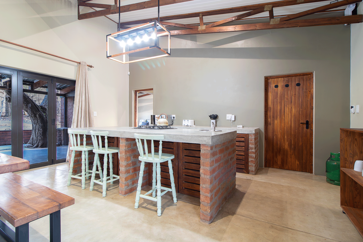 A rustic-style kitchen featuring a large concrete island with a brick base, exposed timber roof trusses, and polished concrete floors, opening to an outdoor area.