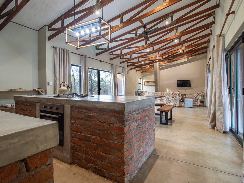 Spacious open-plan living area featuring exposed timber roof trusses, polished concrete floors, and a rustic brick kitchen island with integrated appliances.