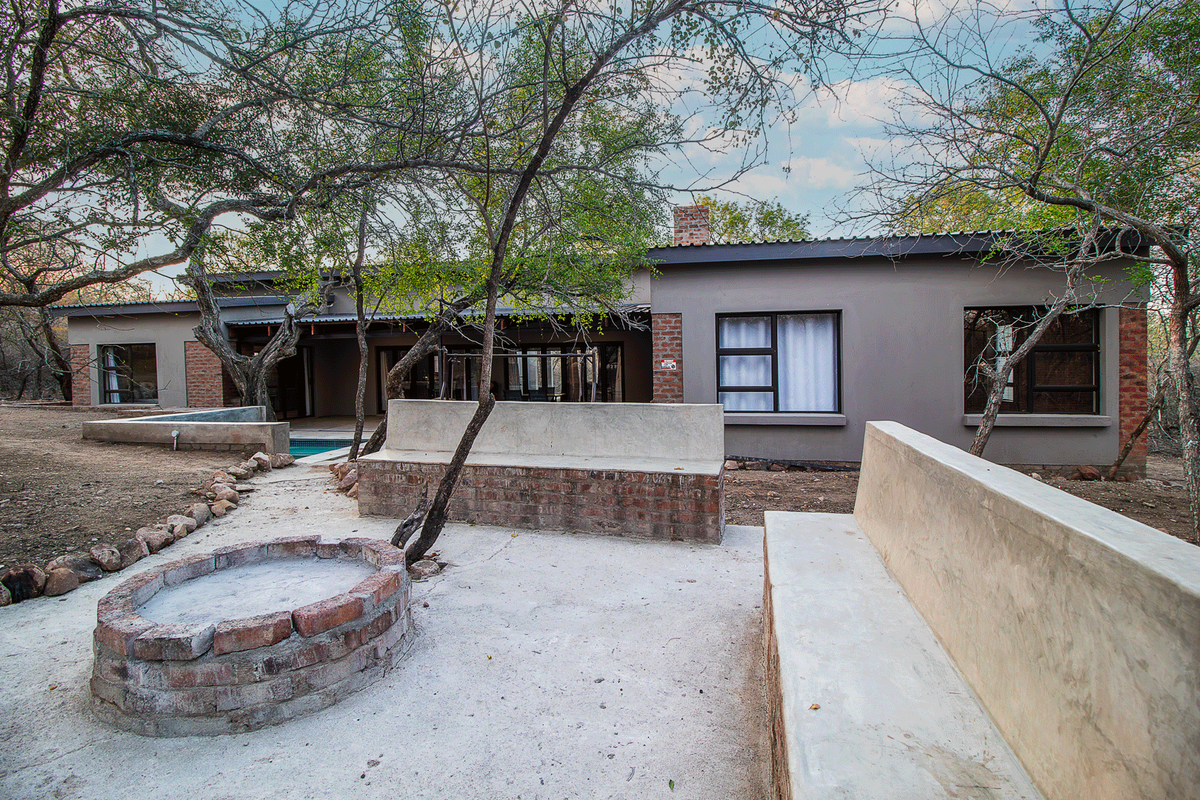 Rear exterior view featuring a built-in brick fire pit, concrete bench seating, and a swimming pool set against a natural bushveld backdrop.