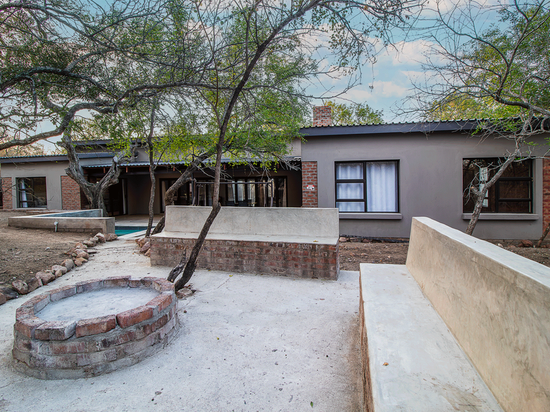 Rear exterior view featuring a built-in brick fire pit, concrete bench seating, and a swimming pool set against a natural bushveld backdrop.