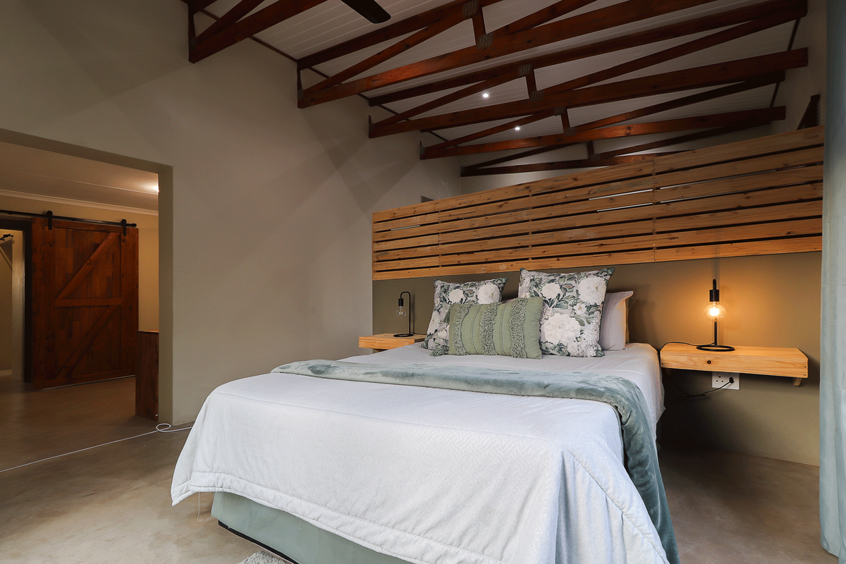 Spacious bedroom featuring exposed timber roof trusses, polished concrete floors, a custom timber slat headboard, and a rustic barn-style sliding door.