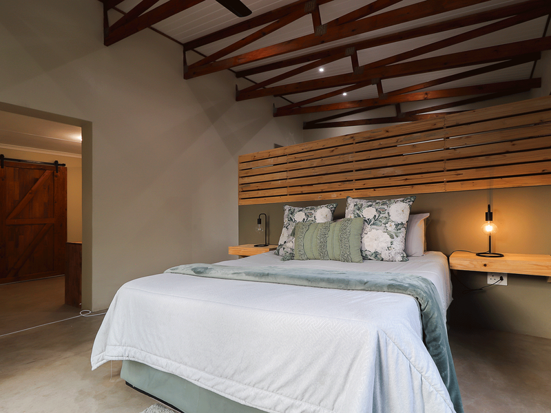 Spacious bedroom featuring exposed timber roof trusses, polished concrete floors, a custom timber slat headboard, and a rustic barn-style sliding door.
