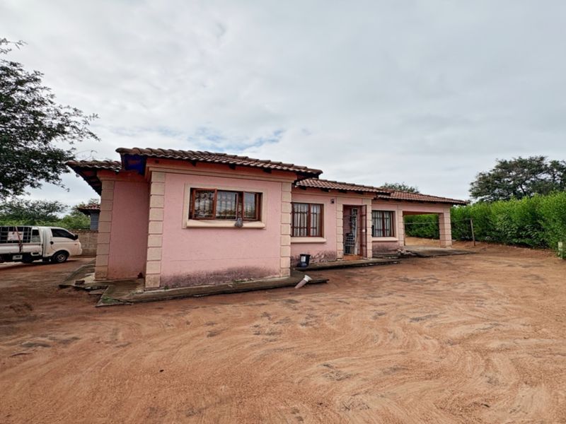 Front exterior view of a single-story house featuring a tiled roof, security bars on windows, and an attached carport on a large dirt lot.