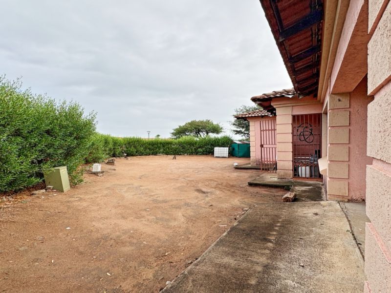 Rear exterior view of the property featuring a concrete walkway, security gate, and a large open yard area with perimeter hedging.