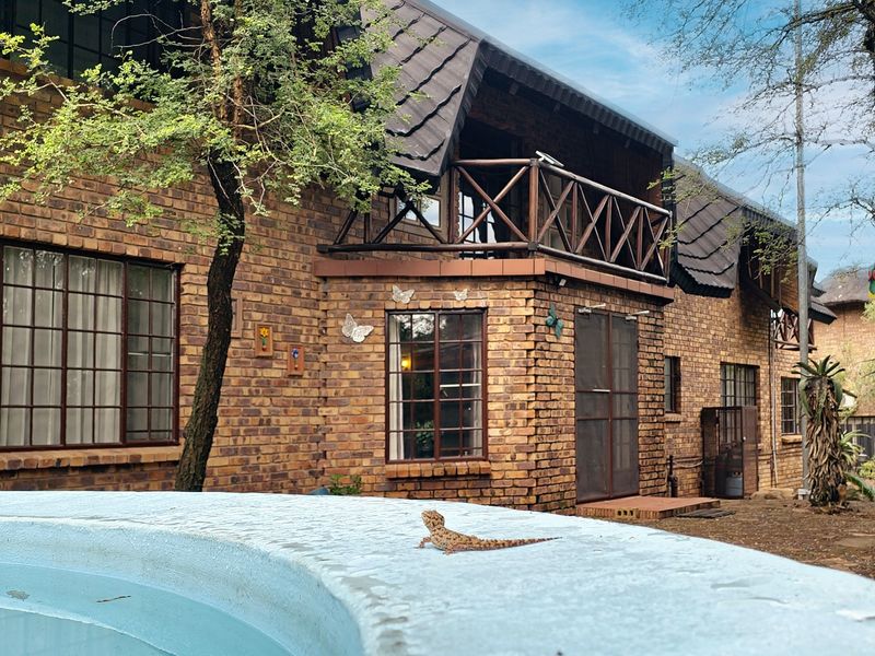 Rear view of a face-brick house featuring a swimming pool, upper-level balcony with wooden railing, and established garden surroundings.