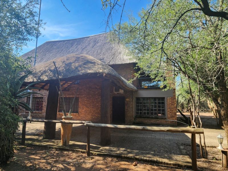 A rustic double-story house featuring a traditional thatch roof, exposed brick exterior, and a covered patio area set within a natural wooded environment.