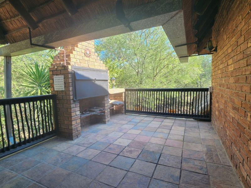 A covered patio featuring a built-in brick braai, tiled flooring, and metal railings, overlooking a natural garden setting.