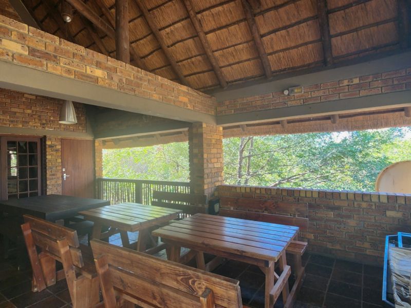 A spacious covered patio featuring exposed brick walls, a traditional thatched roof, and tiled flooring, offering an open-air feel with views of the surrounding trees.