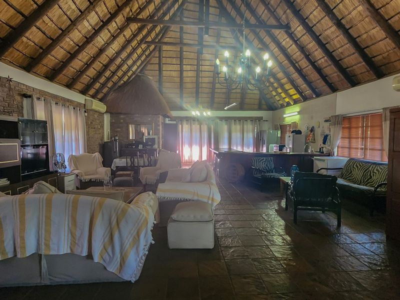 Spacious open-plan living area featuring a high thatched roof with exposed timber trusses, exposed brick walls, tiled flooring, and a built-in bar.