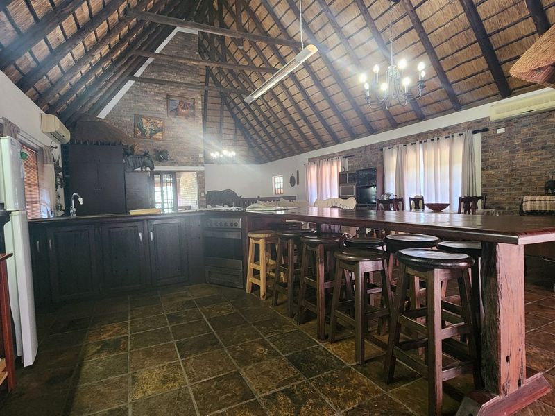 Spacious open-plan kitchen and living area featuring a high thatched ceiling with exposed trusses, rustic brick walls, slate flooring, and a large breakfast bar.