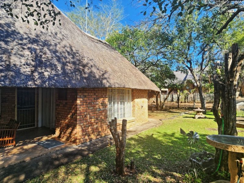 A charming face brick house with a traditional thatch roof, featuring a covered patio overlooking a spacious, tree-lined garden.