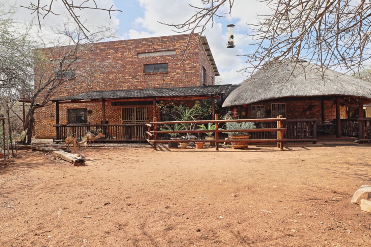 Rear view of a rustic brick house featuring a thatched roof section and a covered timber deck patio area in a natural bushveld setting.