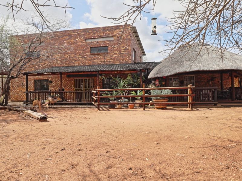 Rear view of a rustic brick house featuring a thatched roof section and a covered timber deck patio area in a natural bushveld setting.