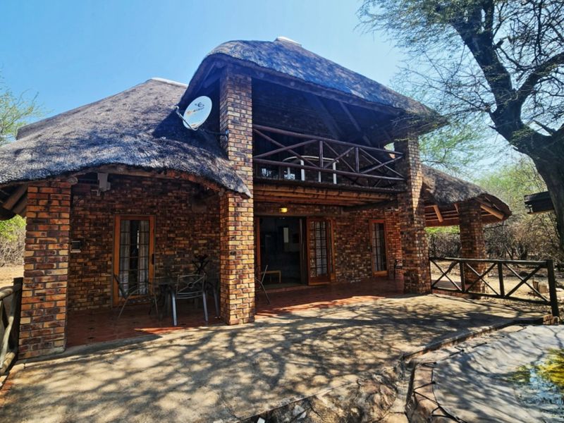 A rustic face brick house featuring a traditional thatched roof, a covered patio area, and an upper-level balcony overlooking the surrounding natural landscape.