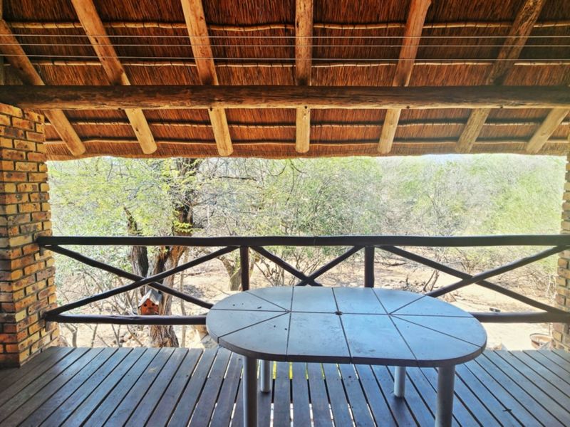 Covered outdoor patio featuring a traditional thatched roof, exposed brick pillars, and timber decking, offering a rustic space with views of the surrounding bushveld.