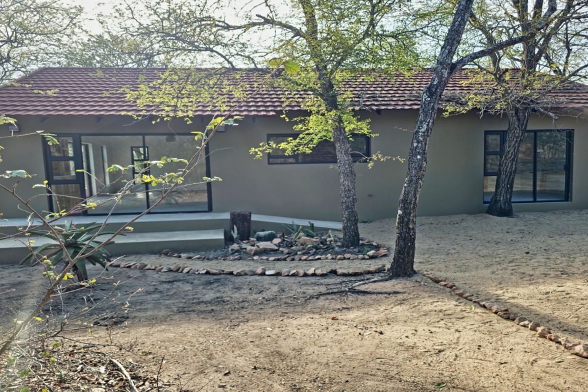 Rear view of a single-story house featuring a tiled roof, sliding glass doors, and a concrete patio set within a natural bushveld garden.