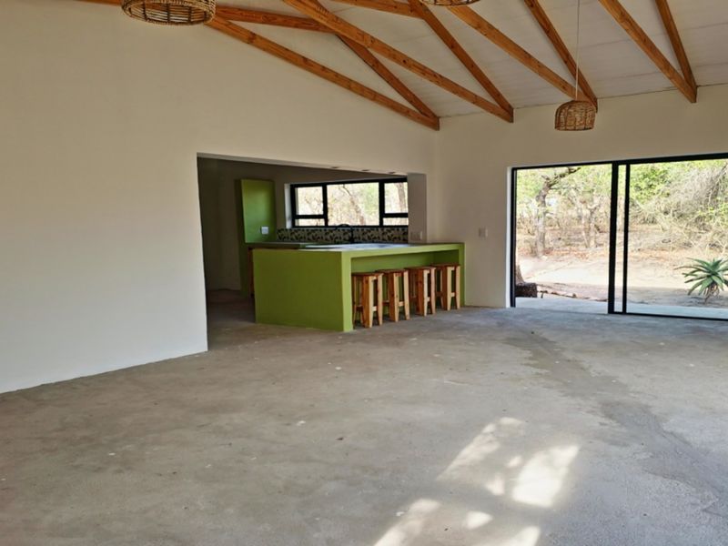 Spacious open-plan living area featuring exposed timber roof trusses, concrete flooring, and a kitchen island, with large sliding doors opening to the outdoors.