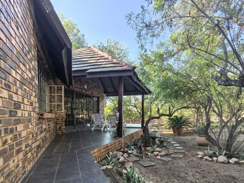 A covered outdoor patio area featuring dark tiled flooring, brick exterior walls, and timber support posts, overlooking a natural garden setting.