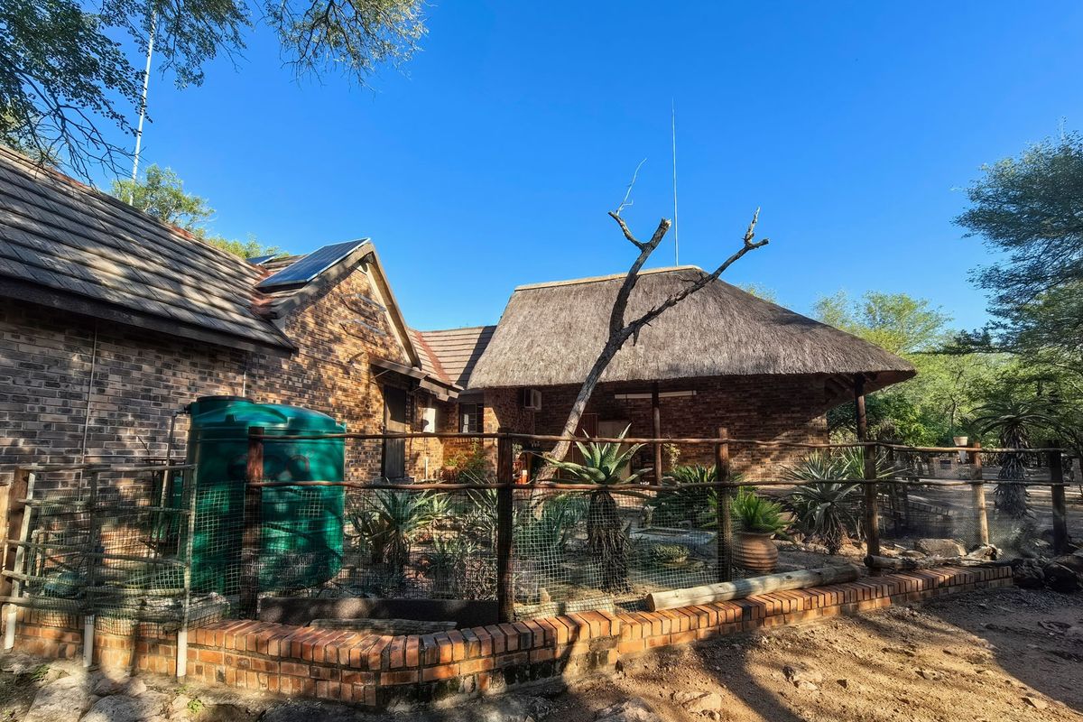 Rear view of a brick house featuring a combination of tiled and thatched roofing, solar panels, and a fenced garden area with a water tank.