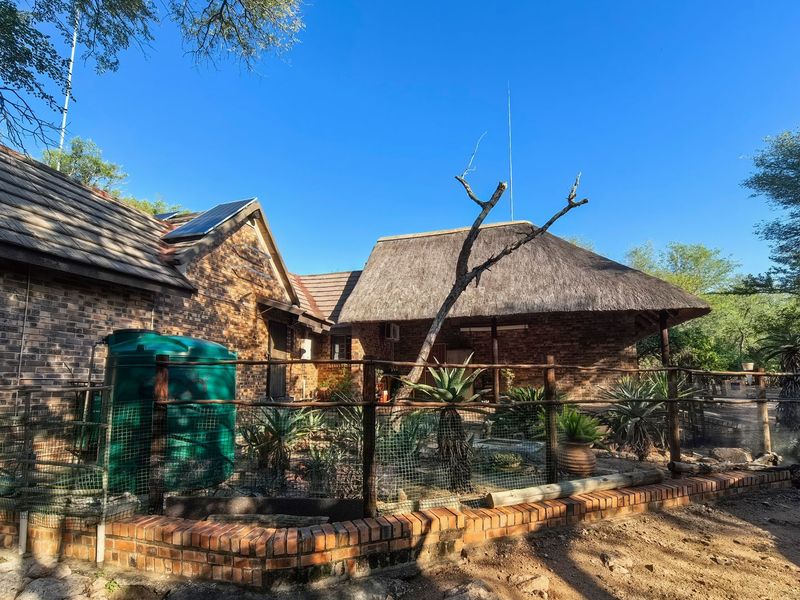 Rear view of a brick house featuring a combination of tiled and thatched roofing, solar panels, and a fenced garden area with a water tank.