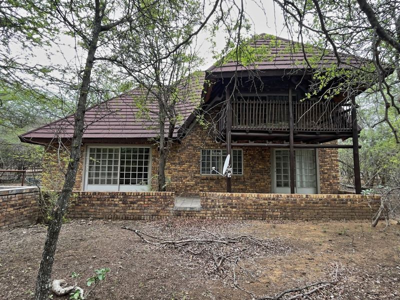 Rear view of a double-story brick house featuring a wooden balcony, sliding glass doors, and a surrounding natural bushveld garden setting.