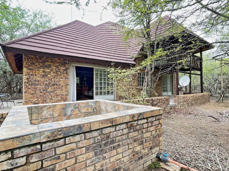 Rear view of a face brick house featuring a raised patio, balcony, and a brick-built water feature set within a natural bushveld environment.