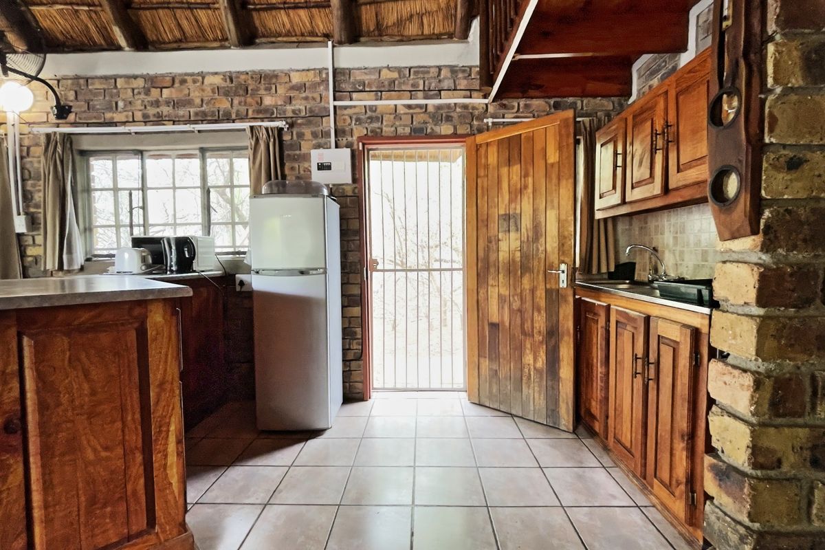 Rustic kitchen featuring exposed brick walls, a traditional thatched ceiling, wooden cabinetry, and tiled flooring with direct access to the exterior.