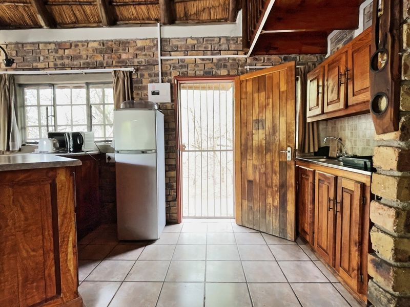 Rustic kitchen featuring exposed brick walls, a traditional thatched ceiling, wooden cabinetry, and tiled flooring with direct access to the exterior.