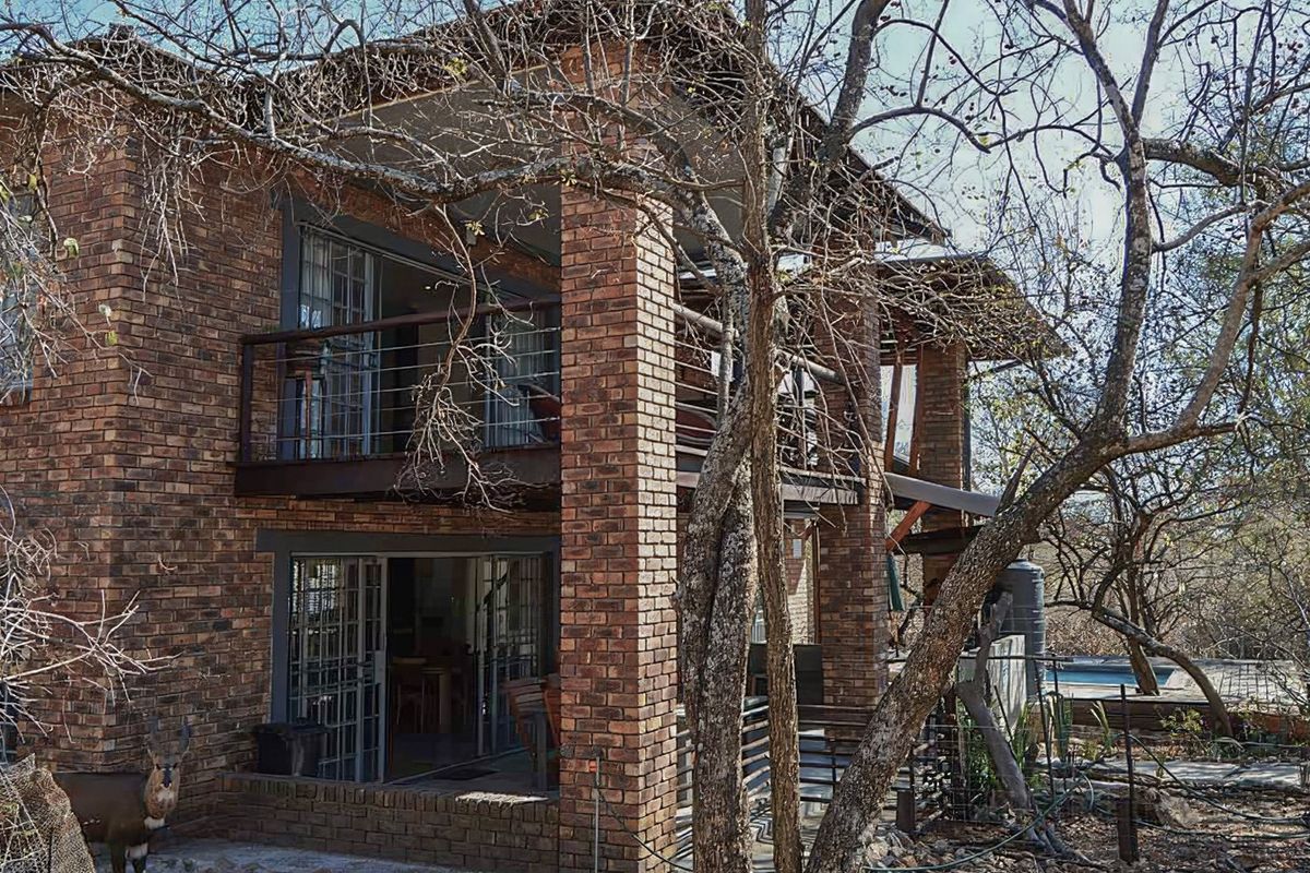 Rear view of a two-story brick house featuring an upper balcony and lower patio with sliding glass doors, set within a natural wooded environment.