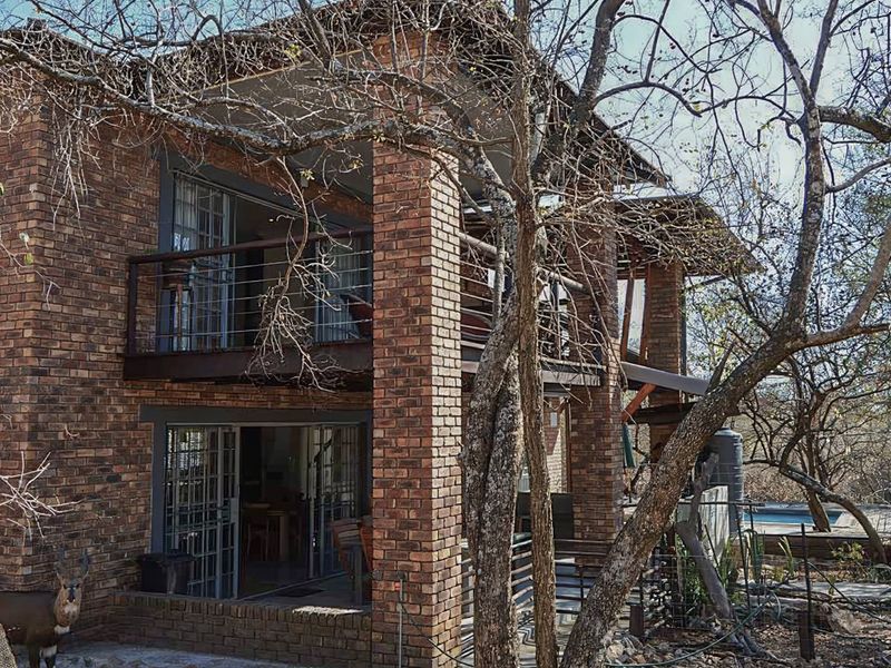 Rear view of a two-story brick house featuring an upper balcony and lower patio with sliding glass doors, set within a natural wooded environment.