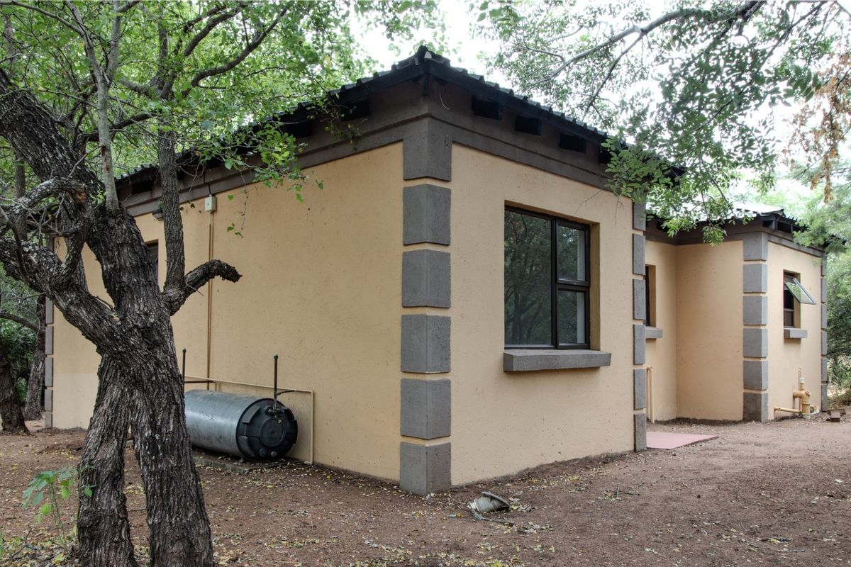 Rear exterior view of the property featuring plastered walls, aluminium window frames, and an external geyser unit set within a wooded garden area.