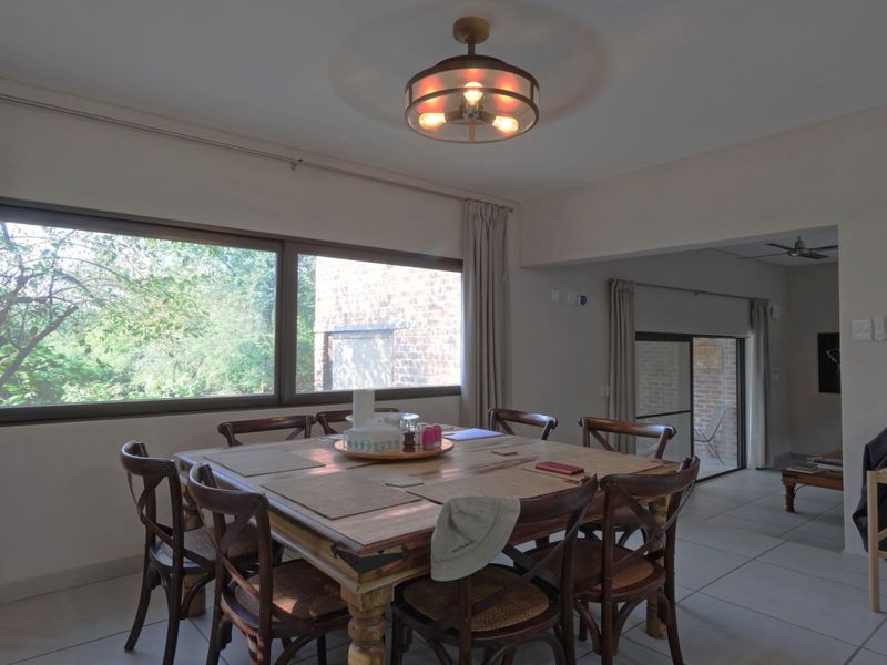 A dining area featuring tiled flooring, a large window providing natural light, and a central ceiling light fixture, with an adjacent open-plan living space.