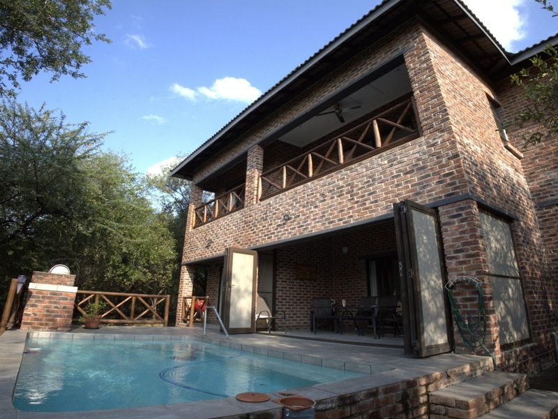 Rear view of a double-story exposed brick house featuring a swimming pool, ground-floor patio, and an upper-level balcony with timber railings.
