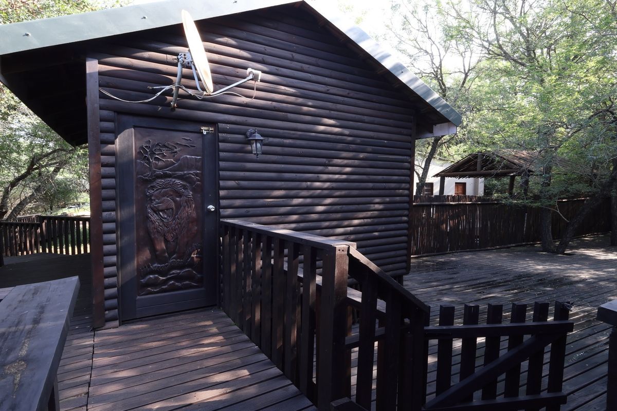 A rustic log cabin exterior featuring a carved timber door, expansive timber decking, and a surrounding wooden balustrade set in a natural wooded environment.