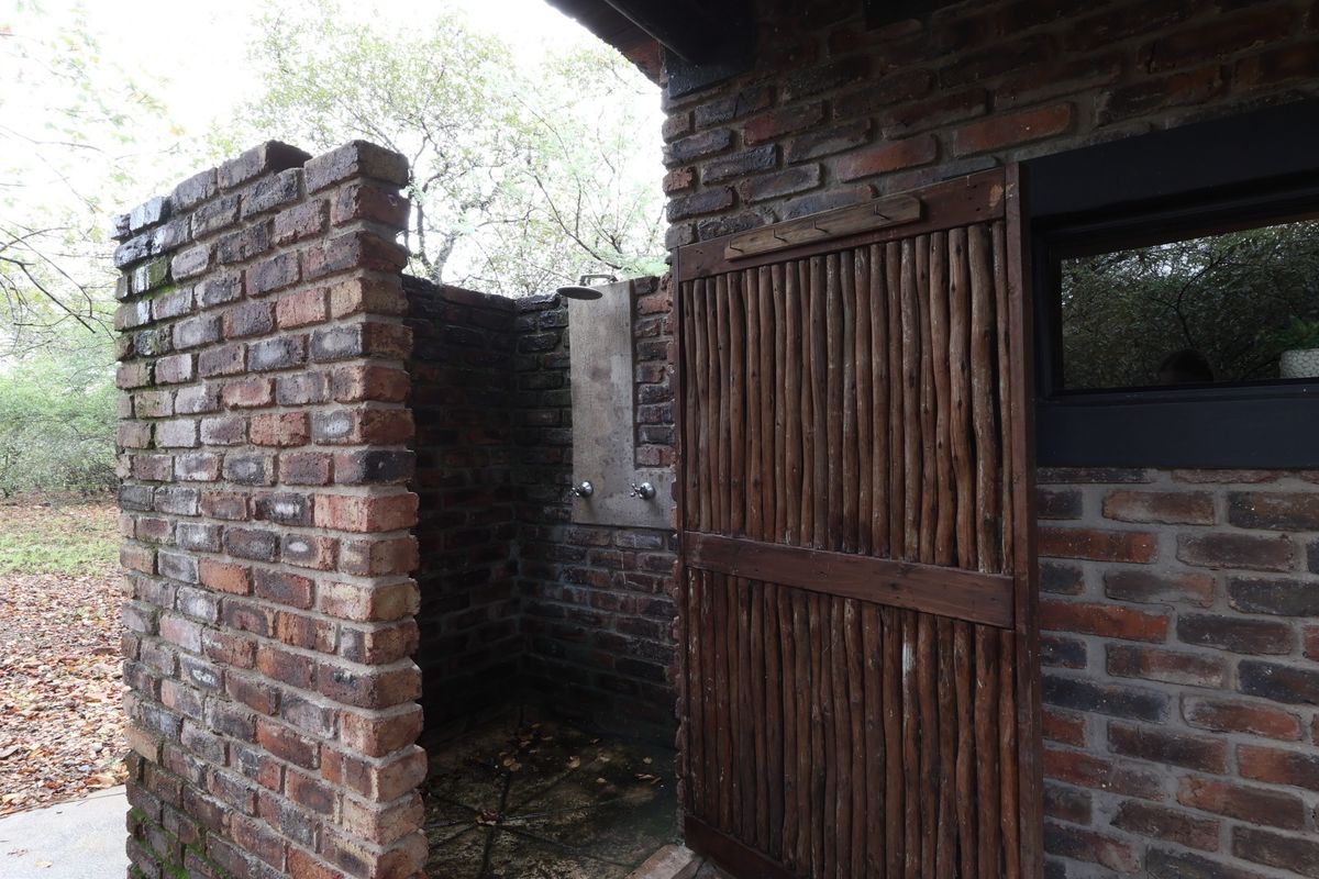 An outdoor shower area featuring exposed brick walls, a timber screen door, and a functional shower fixture set in a natural garden environment.