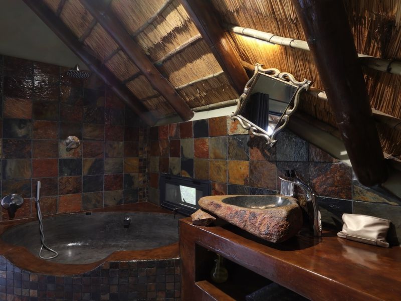 Unique bathroom featuring a stone basin, built-in bathtub, and slate tiling under a rustic thatched roof with exposed timber beams.