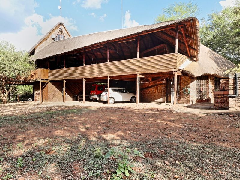 A rustic double-story house featuring a traditional thatch roof, timber balcony, and a spacious covered ground-level parking area.