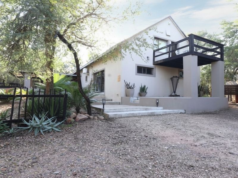 A well-maintained house exterior featuring a raised balcony, concrete entrance steps, and a gravel driveway set within a natural, wooded environment.