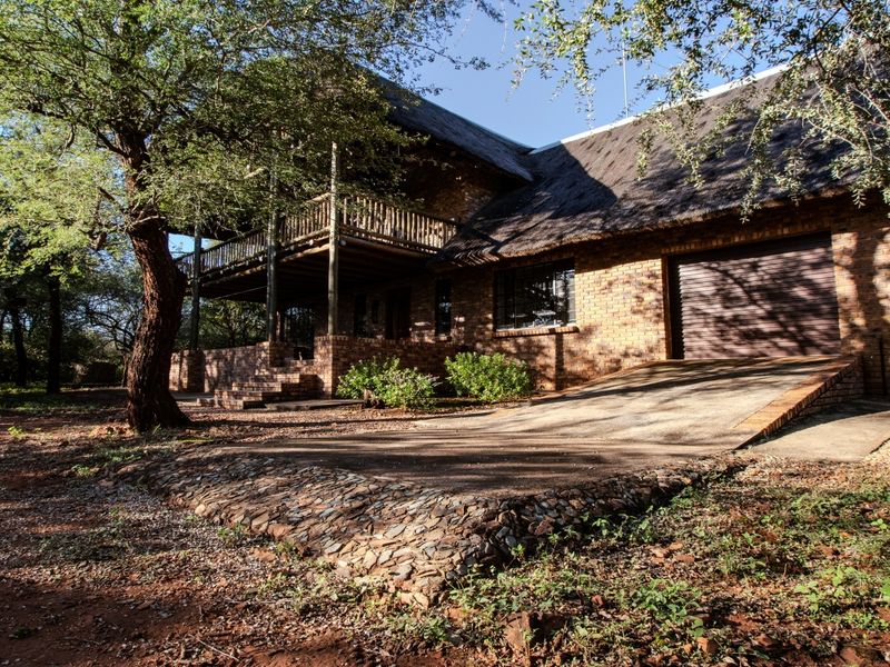 A charming double-story face brick house featuring a traditional thatched roof, a timber balcony, and an integrated garage set within a natural bushveld environment.