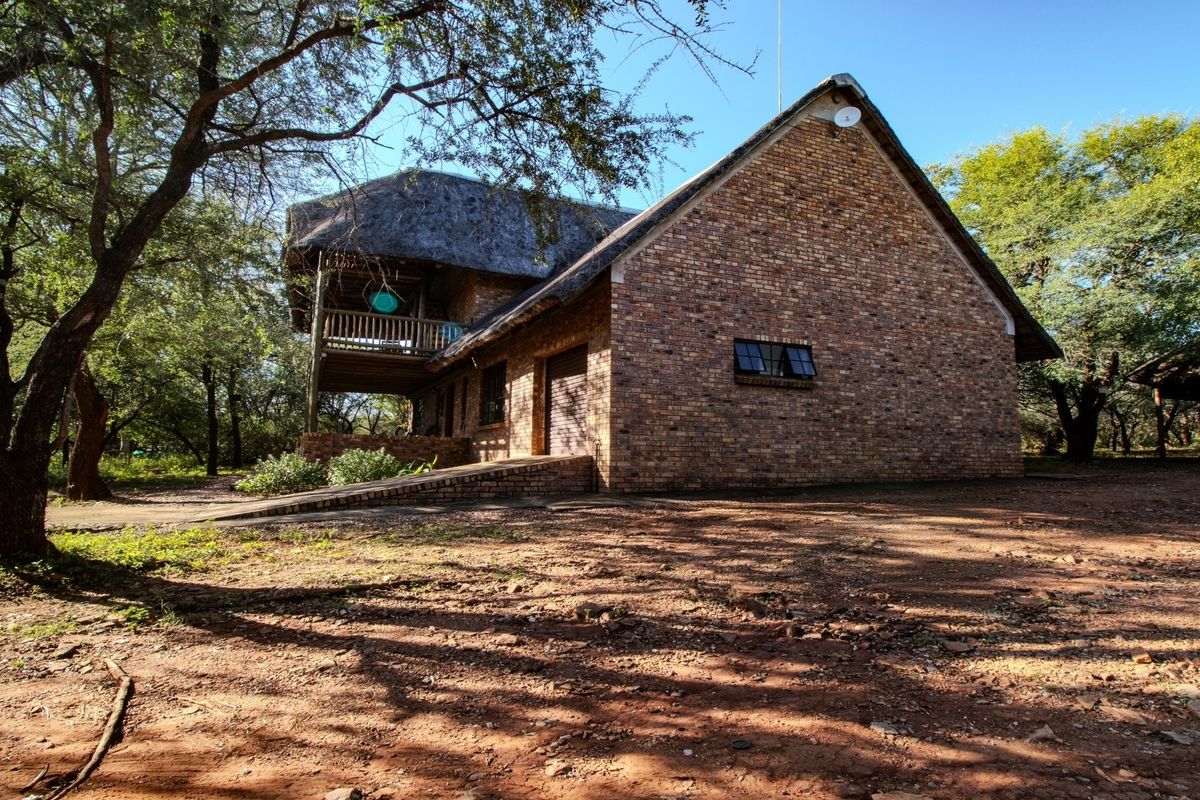 A two-story face brick house featuring a traditional thatched roof, a wooden balcony, and a concrete access ramp, set within a natural bushveld environment.