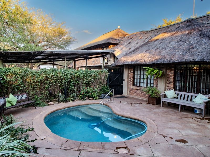 A private outdoor pool area featuring a paved patio, a pergola, and a charming thatched-roof house exterior.